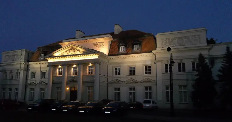 Oświetlona nocą fasada Pałacu Prymasowskiego z kolumnowym portykiem i samochodami na dziedzińcu